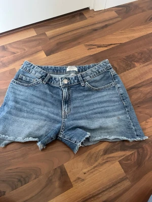 Blå jeansshorts från Lindex - Säljer ett par blå jeansshorts från Lindex i storlek 170. De har klassisk femficksmodell, råa kanter nertill och knappgylf. Perfekta för sommaren och har en avslappnad passform. Materialet är slitstarkt denim i bomull.