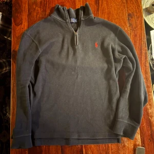 Svart halvzip tröja från Polo Ralph Lauren - Svart långärmad tröja med halv dragkedja och hög krage från Polo Ralph Lauren. Klassisk röd broderad logga på bröstet. Tröjan har ribbade muddar vid ärmslut och nederkant, perfekt för en stilren och avslappnad look. Vintage så har tvättad look där och där