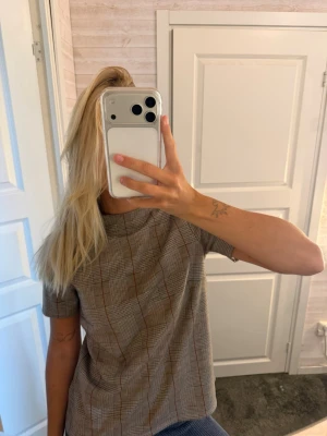 Rutig t-shirt i beige och brunt - Säljer en snygg rutig t-shirt med beige, bruna och svarta toner. T-shirten har korta ärmar och en rak passform. Tyget är mjukt och känns som bomull eller polyester. Perfekt för dig som gillar klassiska mönster med en modern twist.
