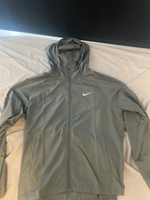 Grå vindjacka med huva från Nike - Snygg grå vindjacka från Nike med huva och hel dragkedja framtill. Jackan har två sidofickor med dragkedja och reflekterande Nike-logga på bröstet. Perfekt för blåsiga dagar och sportiga outfits.