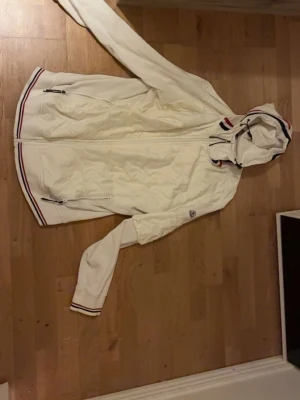 Vit vindjacka med huva från Moncler - Snygg vit vindjacka från Moncler med huva och dragkedja framtill. Jackan har två fickor med dragkedja, Moncler-logga på ärmen och detaljer i rött, vitt och blått vid huvan och nederkanten. Materialet är glansigt och lätt, perfekt för blåsiga dagar.