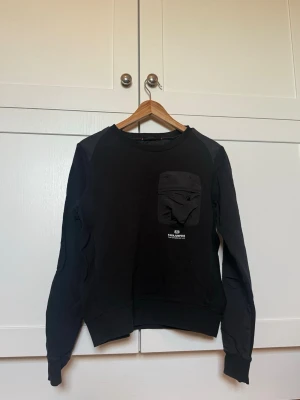 Svart sweatshirt med ficka från Parajumpers - Svart sweatshirt från parajumpers med rund hals och en praktisk ficka på bröstet. Tröjan har långa ärmar och ribbade muddar vid ärmslut och nederkant. Tröjan har ett mer ”jack” tyg på ärmar och ficka. Står storlek Y-L (Youth large) vilket är den största ungdomsstorleken och det står att det är som storleken 16 år. 