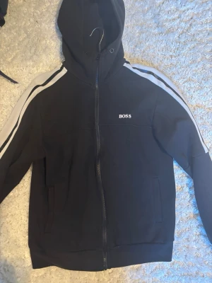 Svart hoodie med dragkedja från BOSS - Svart hoodie från BOSS med vit logga på bröstet och vita ränder längs ärmarna. Modellen har huva och hel dragkedja framtill samt fickor på sidorna. Perfekt för en sportig och avslappnad stil. Storlek S 