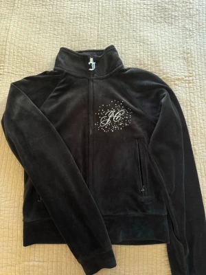 Juicy couture zip - Snygg svart velourtröja med dragkedja och hög krage. På bröstet finns ett JC-märke i strass och dragkedjan har en dekorativ JC-detalj. Tröjan har två fickor med dragkedja och en mjuk, lyxig känsla. På ryggen står det Juicy couture med paljetter. Använd cirka 4 gånger så inga defekter finns.