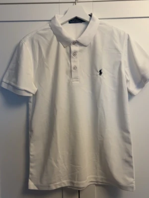 Vit pikétröja från Polo Ralph Lauren - Klassisk vit pikétröja från Polo Ralph Lauren med korta ärmar och krage. Tröjan har knappar framtill och den ikoniska broderade loggan på bröstet. Perfekt till jeans eller shorts för en clean och stilren look.