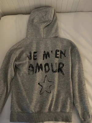 Grå hoodie med tryck och huva - Säljer en grå hoodie med huva och dragsko. På ryggen står det 'JE M'EN AMOUR' i svart text och en stjärna under. Samma tryck finns mindre framtill. Hoodien har ribbade muddar vid ärmar och nederkant. Perfekt för en chill och avslappnad stil.