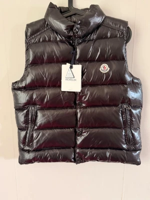 Svart dunväst från Moncler - Snygg svart dunväst från Moncler med glansig finish och klassisk Moncler-logga på bröstet. Västen har hög krage, två dragkedjeförsedda fickor och quiltad design. Perfekt för lager-på-lager och streetstyle.