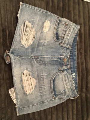 Ljusa blå jeansshorts med slitningar - Säljer ett par ljusa blå jeansshorts med råa kanter och stora slitningar framtill. Shortsen har fem fickor, bälteshällor och knappgylf. Perfekta för en avslappnad och trendig sommarlook.