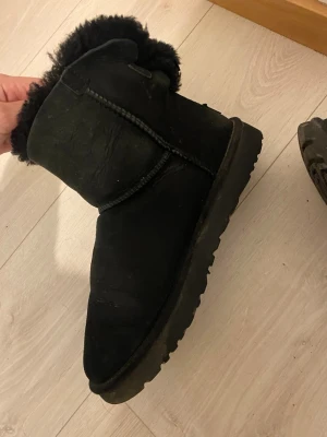 UGGS - Svarta med rosett - Mysiga svarta stövlar i fårskinn med fluffigt foder och dekorativ rosett baktill. Stövlarna har en rundad tå och grov sula som ger bra grepp. Perfekta för kalla dagar när du vill hålla fötterna varma och snygga.