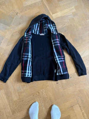 Burberry hoodie  - Säljer en svart hoodie med huva och rutigt foder i rött, vitt och grått på insidan. Hoodien har långa ärmar och ribbade muddar vid ärmslut och nederkant. Perfekt för dig som gillar klassisk stil med en twist. Skriv vid minsta fundering och priset är inte hugget i sten