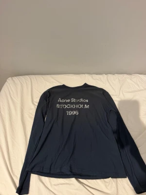 Mörkblå långärmad topp Acne Studios - Mörkblå långärmad topp från Acne Studios med tryck 'Acne Studios STOCKHOLM 1996' på bröstet. Enkel och stilren design, perfekt för dig som gillar minimalistiskt mode. Pris kan diskuteras