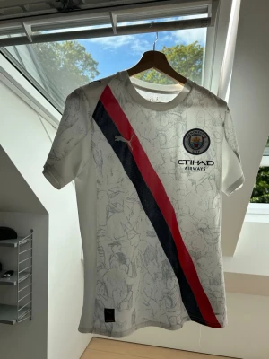Manchester City matchtröja Puma - Vit Manchester City matchtröja från Puma med röd och marinblå diagonal rand på framsidan. Tröjan har klubbmärke och sponsorlogga på bröstet samt subtilt mönster i tyget. Korta ärmar och rund hals. Perfekt för fotbollsfans!