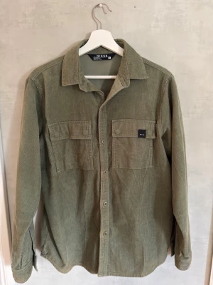 Grön manchester overshirt från Nicce - Snygg overshirt i grön manchester från Nicce. Skjortan har två bröstfickor med lock och knapp, klassisk krage och knäppning framtill. Perfekt lager på lager-plagg med avslappnad passform och lång ärm. Skick 9/10, Storlek S (passar bra till M också), Hör av dig vid några frågor🙌