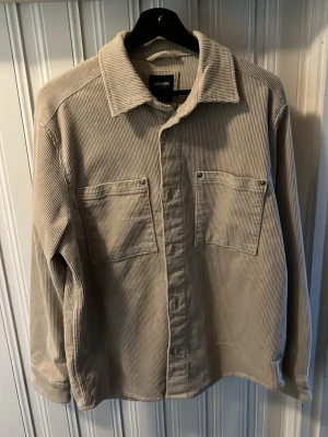 Beige overshirt i manchester - Snygg beige overshirt i manchestertyg med två stora bröstfickor och klassisk krage. Långärmad modell med avslappnad passform, perfekt att slänga över en t-shirt. Stängs med knappar framtill och har en stilren look.