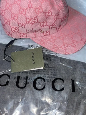 Rosa Gucci keps med GG-mönster - Säljer en rosa keps från Gucci med det klassiska GG-monogrammet över hela kepsen. Kepsen har böjd skärm och justerbart band i brunt läder baktill. Materialet är canvas med exklusiv känsla och detaljer i läder. Perfekt för dig som vill sticka ut med en lyxig accessoar.