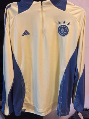 Ajax träningströja gul/blå Adidas - Snygg långärmad träningströja från Ajax i gul och blå färg. Tröjan har halv dragkedja framtill, Ajax klubbmärke och Adidas-logga på bröstet. Tillverkad i 100% polyester, perfekt för träning eller uppvärmning.