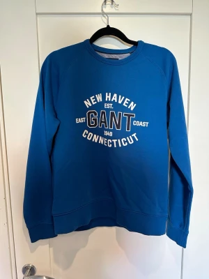 Blå sweatshirt från Gant - Blå sweatshirt från Gant med tryck på bröstet där det står 'New Haven Connecticut'. Tröjan har rund halsringning, långa ärmar och ribbade muddar vid ärmslut och nederkant. Perfekt för en avslappnad och sportig stil.