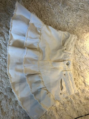 Vit volangkjol från Perfect Jeans - Supersöt vit kortkjol från Perfect Jeans med volangdetaljer och råa kanter. Kjolen har klassisk jeanslook med fickor fram och knappstängning. Perfekt för dig som vill ha en trendig och lekfull stil till sommaren.