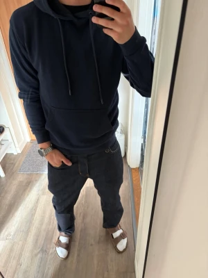 Mörkblå hoodie med huva - Säljer en stilren mörkblå hoodie med huva och dragsnören. Hoodien har en stor magficka framtill och långa ärmar. Perfekt för dig som gillar en enkel och clean look. Materialet känns mjukt och bekvämt.
