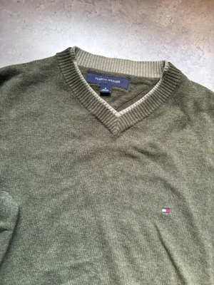 Olivgrön stickad tröja Tommy Hilfiger - Olivgrön stickad tröja från Tommy Hilfiger med V-ringning och ribbade muddar. Tröjan har en liten broderad logga på bröstet och kontrastfärg vid halsringningen. Perfekt för dig som gillar klassisk och stilren design.