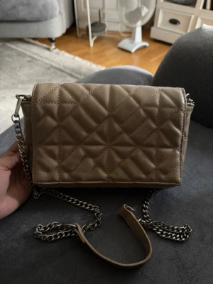 Beige quiltad handväska med kedja - Snygg beige handväska med quiltat mönster och silverfärgad kedjerem. Väskan har magnetlås och en stilren, rektangulär form. Perfekt för dig som gillar trendiga accessoarer med en clean look. Materialet ser ut att vara konstläder.