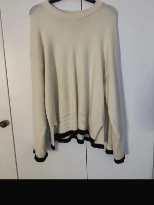 Oversized cremevit stickad tröja - Säljer en oversized cremevit stickad tröja med svarta ribbade detaljer vid ärmslut och nederkant. Tröjan har rund halsringning och slitsar på sidorna för en avslappnad look. Perfekt för lager på lager och enkel att styla.