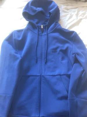 Blå zip hoodie med huva - Snygg blå hoodie med dragkedja och huva. Hoodien har dragsnören i halsen och två stora fickor framtill. Liten diskret logga på bröstet. Perfekt för en avslappnad och sportig stil.