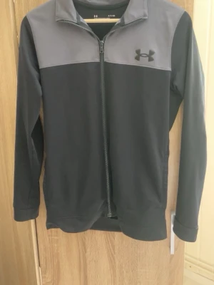 Svart och grå träningsjacka Under Armour - Snygg svart och grå träningsjacka från Under Armour med hel dragkedja framtill och hög krage. Jackan har lång ärm och logga på bröstet. Perfekt för träning eller uppvärmning.