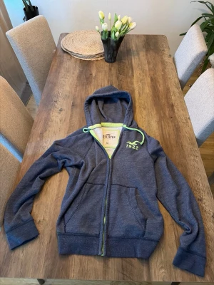 Vintage hollister hoodie  - Mörkblå hoodie från Hollister med dragkedja framtill och två fickor. Grön logga och text '1922' på bröstet samt gröna detaljer på snöret i huvan. Tillverkad i mjukt bomullsmaterial, perfekt för en avslappnad stil.