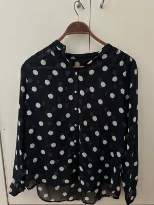 Svart transparent prickig blus - Använd ett par gånger. Snygg svart blus i transparent material med stora vita prickar och knäppning framtill. Blusen har långa ärmar och en avslappnad passform, perfekt för en trendig och lekfull look. Materialet känns lätt och luftigt.