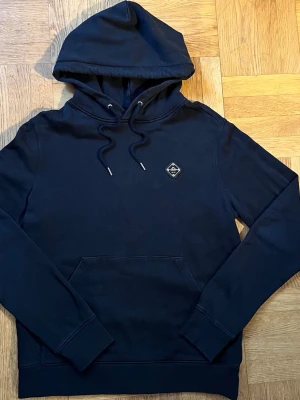 Svart J.Lindeberg hoodie med märke på bröstet  - Snygg svart  hoodie med huva och dragsnören. Har en praktisk magficka och en liten svart patch med vitt tryck på bröstet. Tillverkad i mjukt bomullstyg och har ribbade muddar vid ärmar och nederkant. Perfekt för en avslappnad stil.