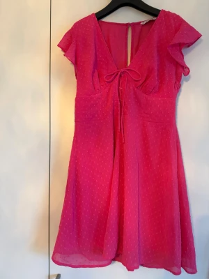 Rosa kortklänning med knyt och volang - Söt rosa kortklänning med v-ringning, knyt framtill och volangärmar. Klänningen har ett diskret prickigt mönster och är tillverkad i ett lätt, luftigt material som ger ett flowy fall. Perfekt för varma dagar och somriga tillfällen. Upplevs liten i storleken.