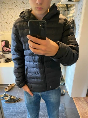 Svart dunjacka med huva - Säljer en svart dunjacka med huva och dragkedja framtill. Jackan har en quiltad design med horisontella sömmar och två sidofickor. Perfekt för kalla vinterdagar och riktigt skön att bära till jeans eller andra vardagskläder.