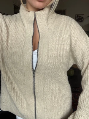 Beige stickad kofta med dragkedja - Mysig beige stickad kofta med hög krage och grov ribbstickning. Koftan har en hel dragkedja framtill och långa ärmar med breda muddar. Perfekt för lager på lager och enkel att matcha med olika outfits.