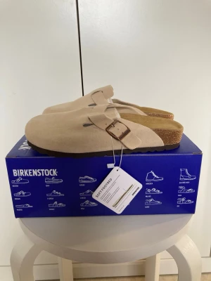  Birkenstock Boston - 37 - Säljer ett par beige Birkenstock Boston loafers i mocka med justerbart spänne i metall. Skorna har en klassisk slip-on design och mjuk fotbädd i kork för extra komfort. Perfekta för dig som gillar stilrena och bekväma skor. Storlek 37 helt nya . 