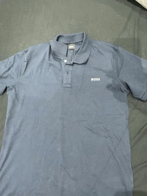 Blå pikétröja från BOSS - Klassisk blå pikétröja från BOSS med krage och knappar framtill. Tröjan har korta ärmar och diskret BOSS-logga broderad på bröstet. Perfekt för en stilren och avslappnad look.