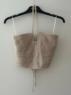 Beige virkad korsettopp med snörning - Trendig beige virkad korsettopp med snörning både fram och bak. Toppen är ärmlös och har tunna axelband samt halterneck-knytning. Perfekt för sommaren och ger en bohemisk vibe till din outfit🌟 Aldrig använd därav prislappen
