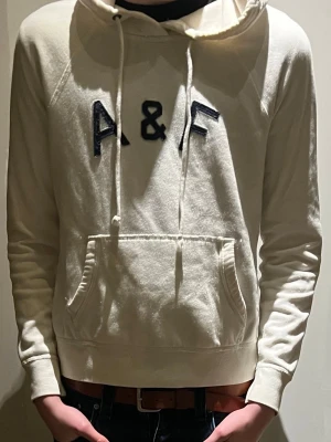 Vit hoodie från Abercrombie & Fitch xs/xss - Snygg vit hoodie från Abercrombie & Fitch med broderad A&F-logga på bröstet. I extremt fint tygg. Hoodien har huva med dragsko och en stor magficka. Perfekt för en chill och avslappnad stil. Mjuk bomullskänsla och klassisk passform. Små fläckar på ryggen men syns knappt vid användning. Därför det låga priset liten i storlek