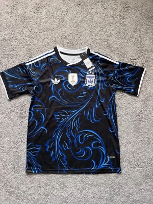 Argentina away  Adidas svart-blå matchtröja - Snygg svart away matchtröja från Adidas med Argentinas landslagsmärke, FIFA-märke och blått bladmönster. Tröjan har vita detaljer på krage och ärmslut samt klassiska Adidas-ränder på axlarna. Tillverkad i lätt och ventilerande Climacool-material.