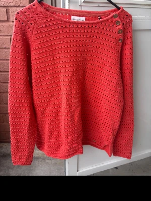 Korallröd stickad tröja med knappar - Snygg korallröd stickad tröja med hålmönster och dekorativa knappar på ena axeln. Tröjan har rund halsringning och långa ärmar. Perfekt för lager-på-lager och ger en färgklick till din outfit.