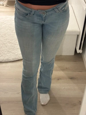 Ljusblå bootcut jeans med brodyr - Säljer ett par ljusblå bootcut jeans med snygg passform och broderade detaljer i rött på bakfickorna. Jeansen har klassisk femficksdesign och är tillverkade i ett mjukt denimtyg med lite stretch för extra komfort. Superliten slitning på höger bakficka