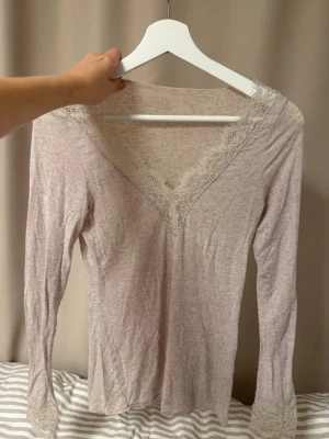 Beige långärmad intimisssmi - Supersöt ljusrosa långärmad topp i mjukt material med spetsdetaljer vid v-ringningen och ärmsluten. Perfekt för mysiga hemmakvällar eller som ett extra lager. Tunn och stretchig för maximal komfort.