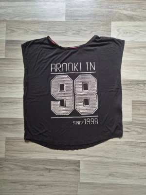 Svart t-shirt med tryck 98 - Svart t-shirt med stort vitt tryck på framsidan där det står 'Rookie 98' och 'Since 1998'. T-shirten har korta ärmar och en lös passform. Perfekt för dig som gillar sportig och avslappnad stil.