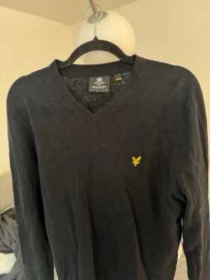 Svart stickad tröja från Lyle & Scott - Klassisk svart stickad tröja från Lyle & Scott med V-ringning och den ikoniska gula örnloggan broderad på bröstet. Tröjan har lång ärm och är tillverkad i mjukt material som passar perfekt till lager på lager-stil.