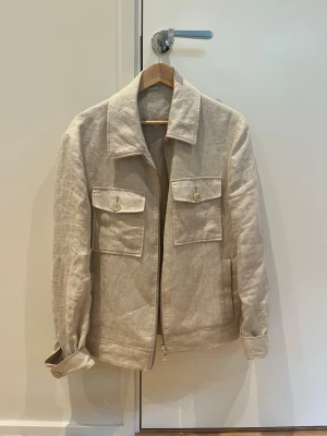 Beige linnéjacka från These Glory Days - Stilren beige jacka i linne från These Glory Days. Jackan har två stora bröstfickor med knappar, klassisk krage och dragkedja framtill. Perfekt för dig som gillar en enkel och clean look.