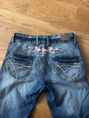 Crazy age jeans med broderi och detaljer - Säljer ett par lågmidjade jeans från Crazy Age med snygga broderade detaljer på bakfickorna i rosa och grönt samt dekorativa sömmar fram. Ca 34cm rakt över midjan 💕