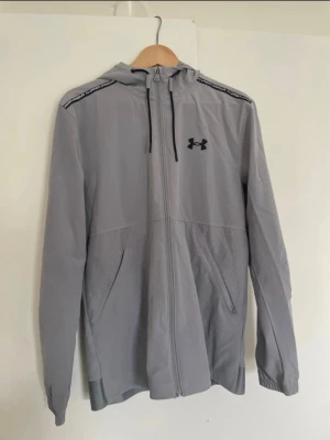 Grå vindjacka från Under Armour - Snygg grå vindjacka från Under Armour med hel dragkedja, huva och svarta detaljer. Jackan har två sidofickor, meshpaneler för ventilation och elastiska muddar vid ärmsluten. Perfekt för blåsiga dagar och sportiga outfits.