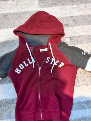 Vintage Vinröd hoodie från Hollister - Snygg vinröd hoodie från Hollister med dragkedja, vita snören och grå ärmar med vit detalj. Stor logga på bröstet och fickor framtill. Perfekt för en avslappnad och trendig stil. Storlek S