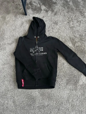 Svart hoodie från Alpha Industries - Svart hoodie med dragkedja från Alpha Industries. Tryck på bröstet med logga och text, samt två fickor framtill. Huva med snörning och metallöljetter. Perfekt för en avslappnad streetwear-look.