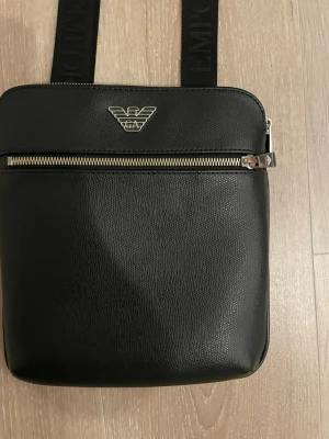 Svart axelväska från Emporio Armani - Snygg svart axelväska från Emporio Armani i konstläder med silverfärgad dragkedja och logga framtill. Justerbar axelrem med diskret logotyptext. Perfekt för att bära det viktigaste med stil.